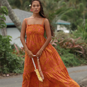 woman with brown hair wearing an orange long maxi dress with smocked bodice and spaghetti straps with puakenikeni print