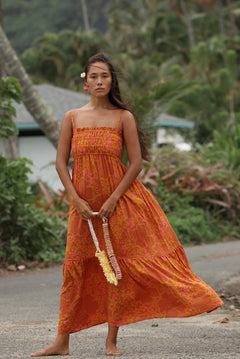 woman with brown hair wearing an orange long maxi dress with smocked bodice and spaghetti straps with puakenikeni print
