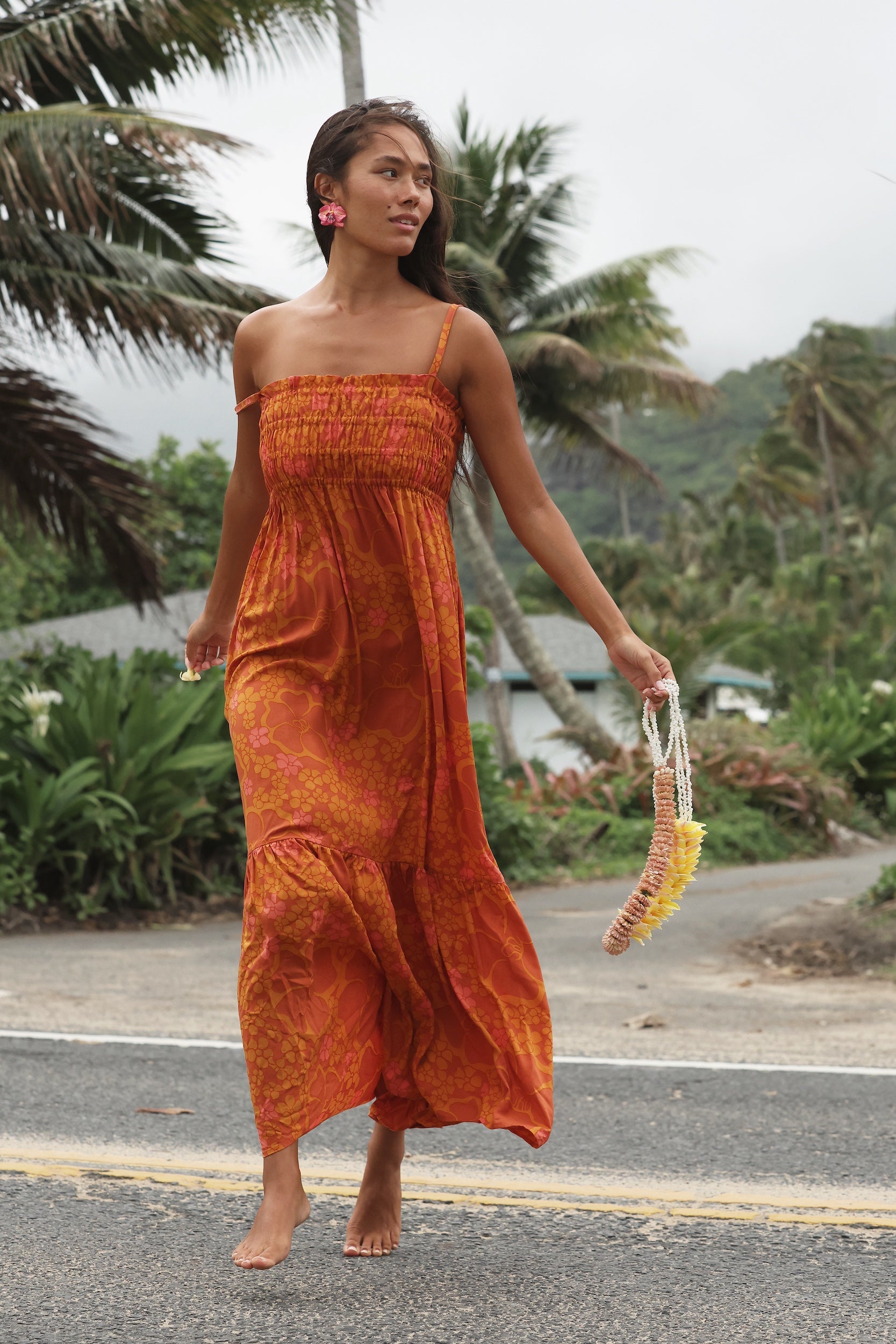 woman with brown hair wearing an orange long maxi dress with smocked bodice and spaghetti straps with puakenikeni print
