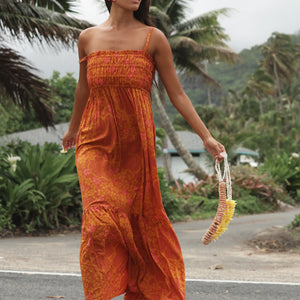 woman with brown hair wearing an orange long maxi dress with smocked bodice and spaghetti straps with puakenikeni print