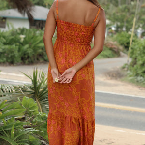 woman with brown hair wearing an orange long maxi dress with smocked bodice and spaghetti straps with puakenikeni print