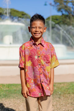 little boy wearing terracotta colored aloha shirt with large purple and yellow flower print 