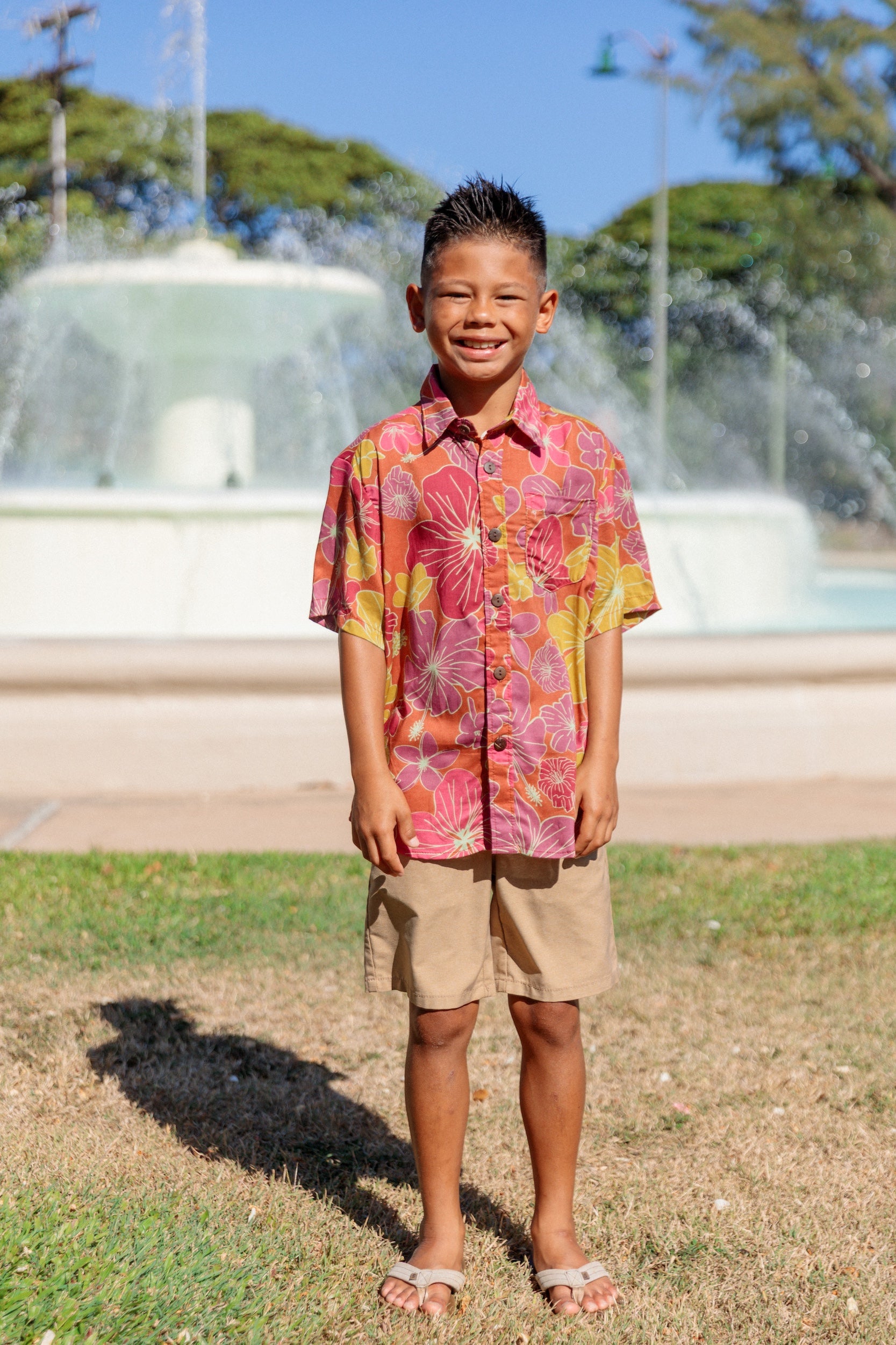 little boy wearing terracotta colored aloha shirt with large purple and yellow flower print 