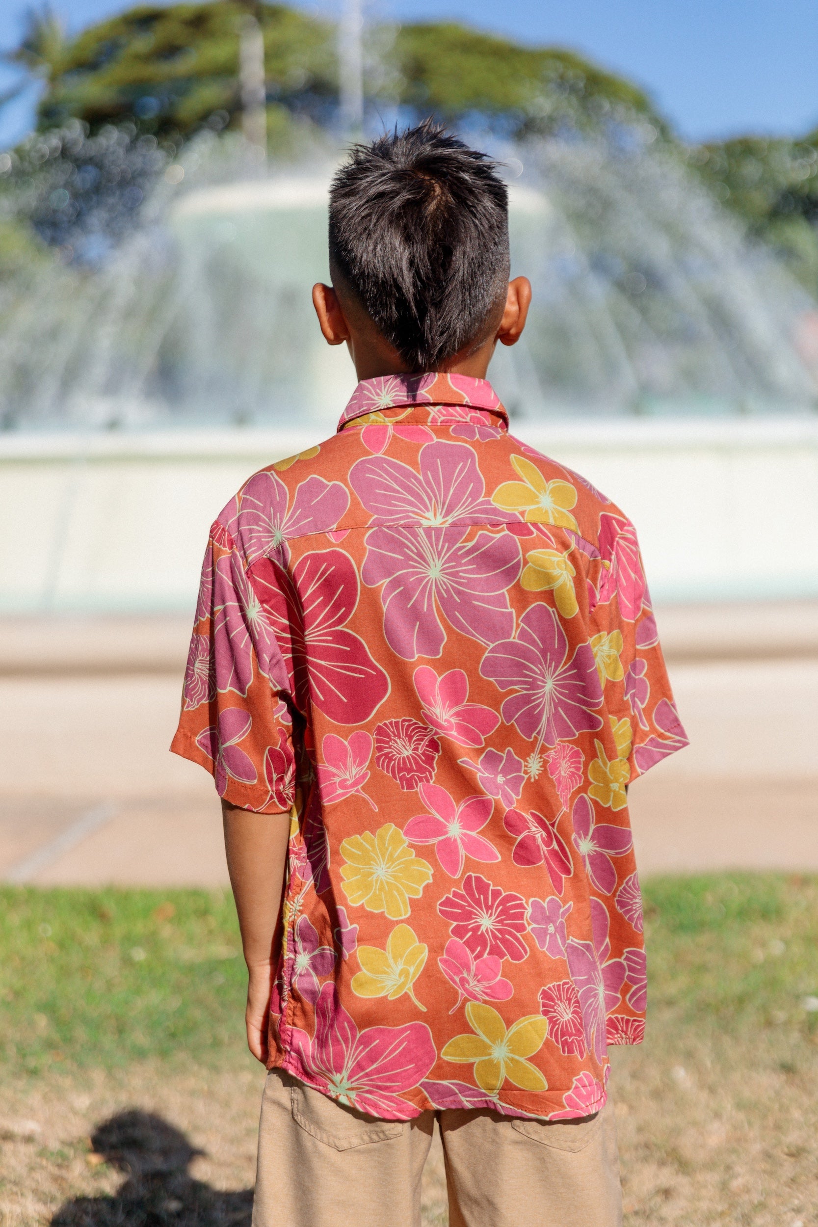 little boy wearing terracotta colored aloha shirt with large purple and yellow flower print 
