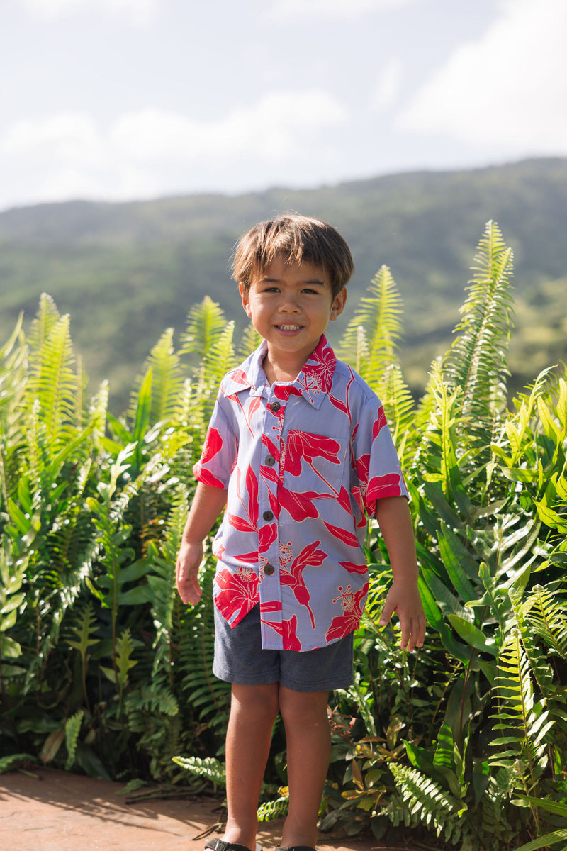 little boy with brown hair wearing a button up aloha shirt with periwinkle background and bright red hibiscus print.
