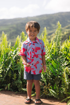 little boy with brown hair wearing a button up aloha shirt with periwinkle background and bright red hibiscus print.