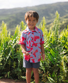 little boy with brown hair wearing a button up aloha shirt with periwinkle background and bright red hibiscus print.
