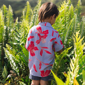 little boy with brown hair wearing a button up aloha shirt with periwinkle background and bright red hibiscus print.