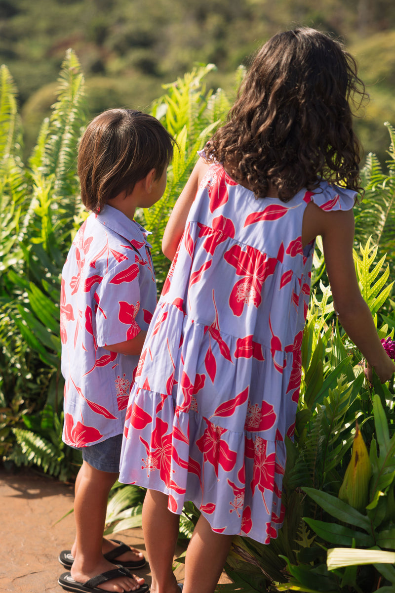 little boy with brown hair wearing a button up aloha shirt with periwinkle background and bright red hibiscus print.