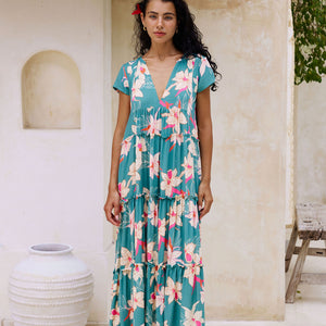 woman with brown hair wearing a tiered maxi dress in teal with large cream colored orchids.