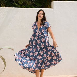 woman with black hair wearing a tiered maxi dress with v neckline in a navy and pink hibiscus print. 