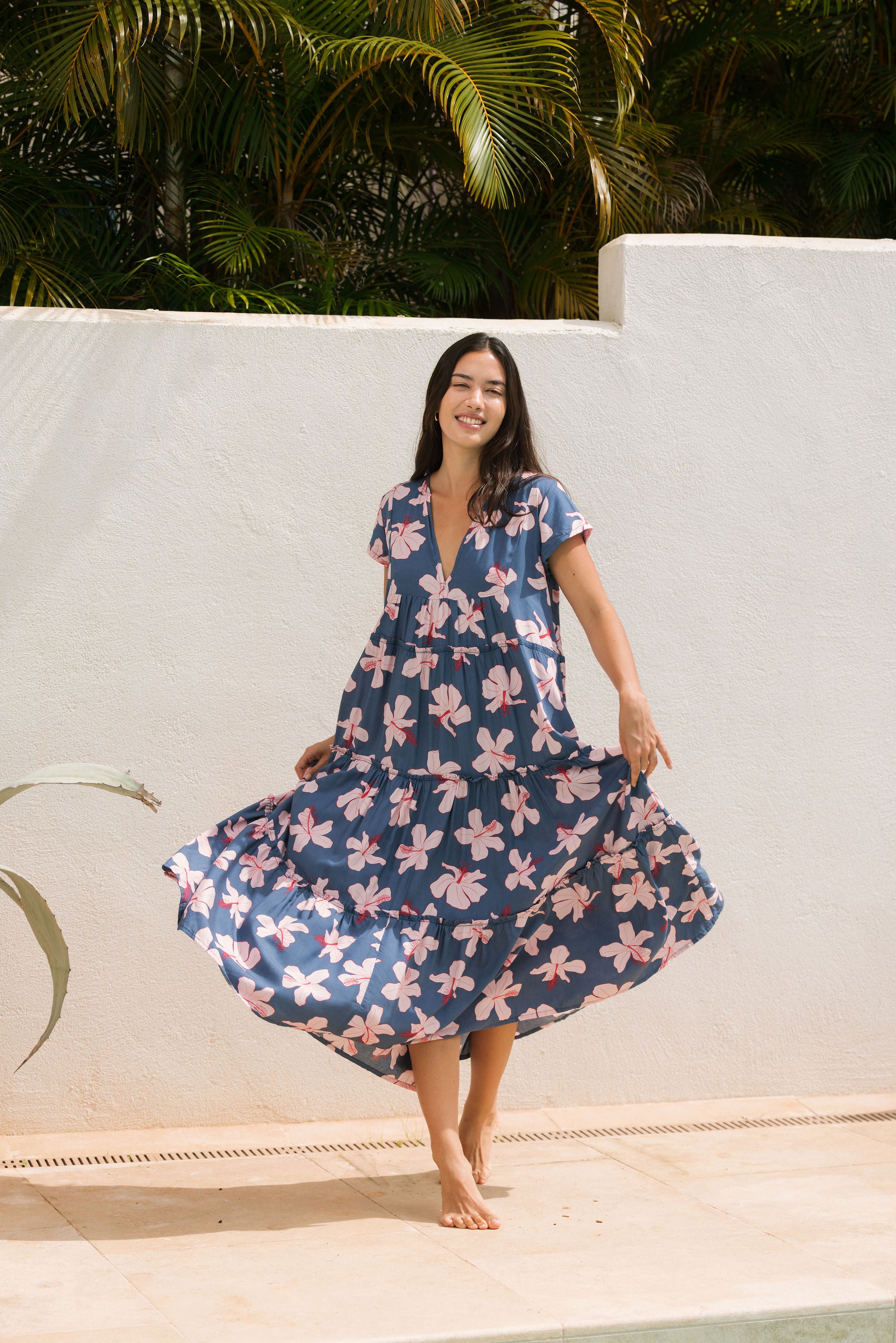 woman with black hair wearing a tiered maxi dress with v neckline in a navy and pink hibiscus print. 