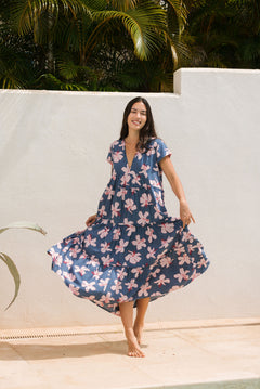woman with black hair wearing a tiered maxi dress with v neckline in a navy and pink hibiscus print. 