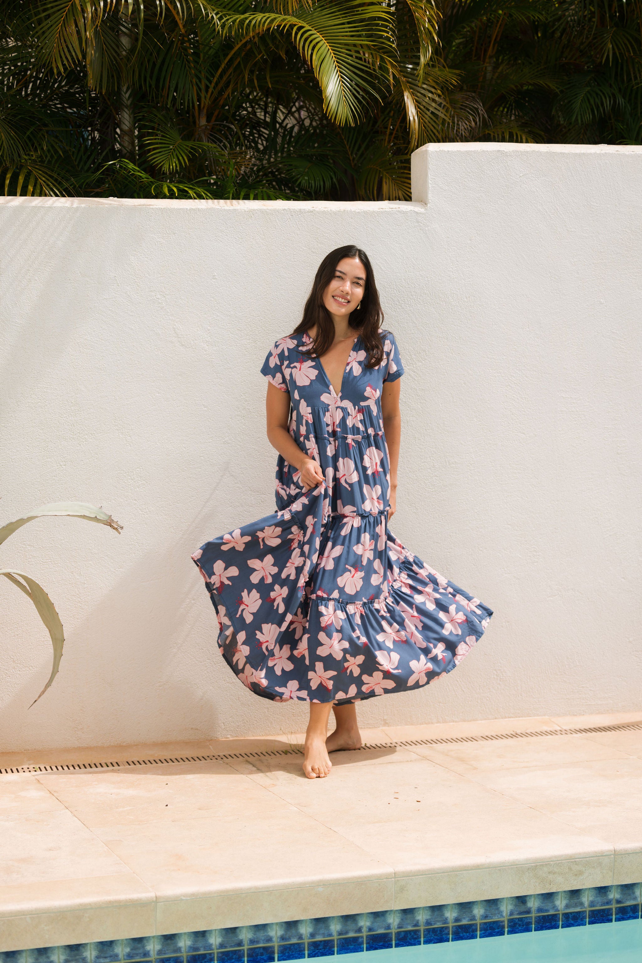 woman with black hair wearing a tiered maxi dress with v neckline in a navy and pink hibiscus print. 