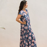 woman with black hair wearing a tiered maxi dress with v neckline in a navy and pink hibiscus print. 