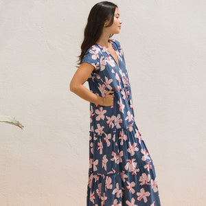 woman with black hair wearing a tiered maxi dress with v neckline in a navy and pink hibiscus print. 