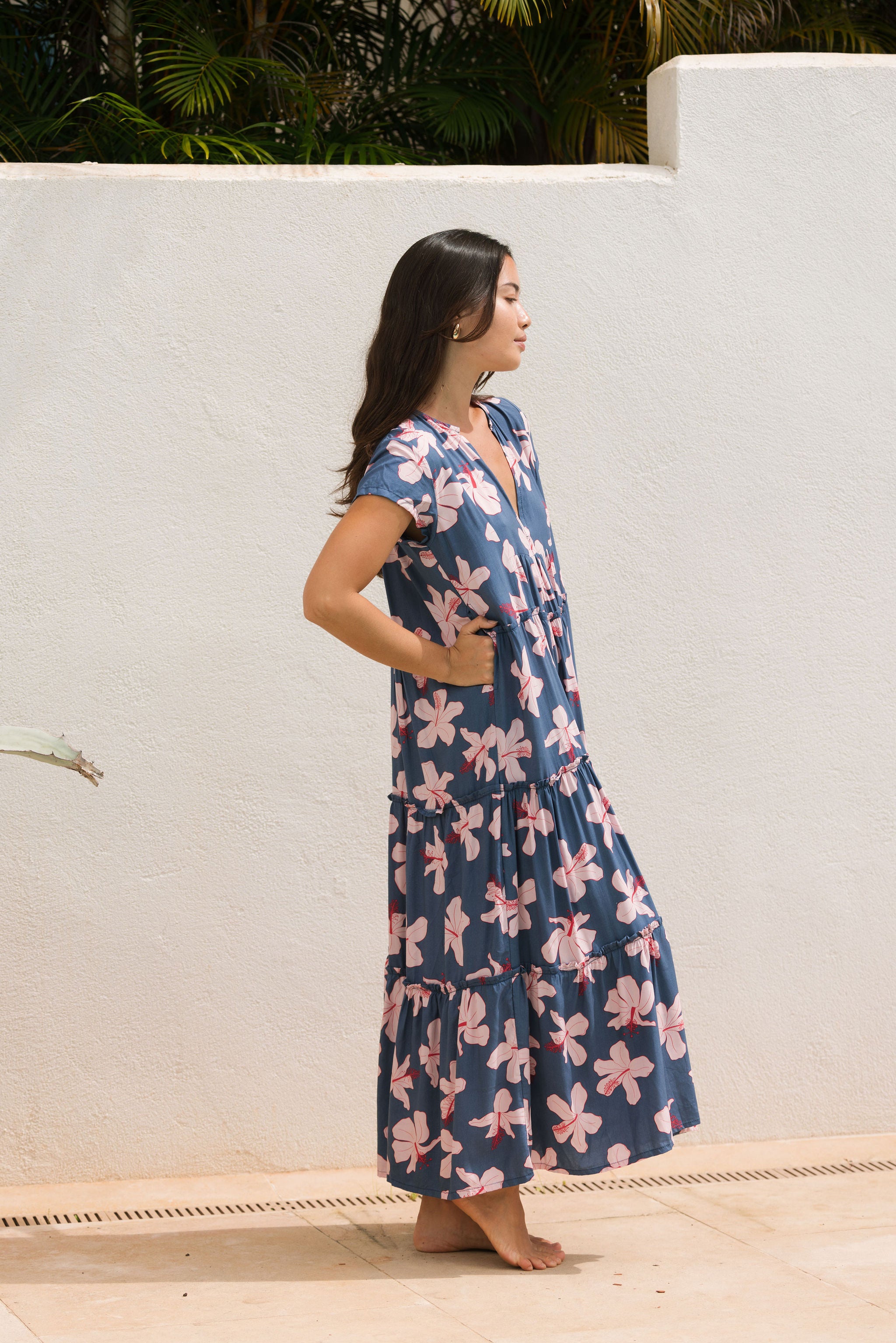 woman with black hair wearing a tiered maxi dress with v neckline in a navy and pink hibiscus print. 