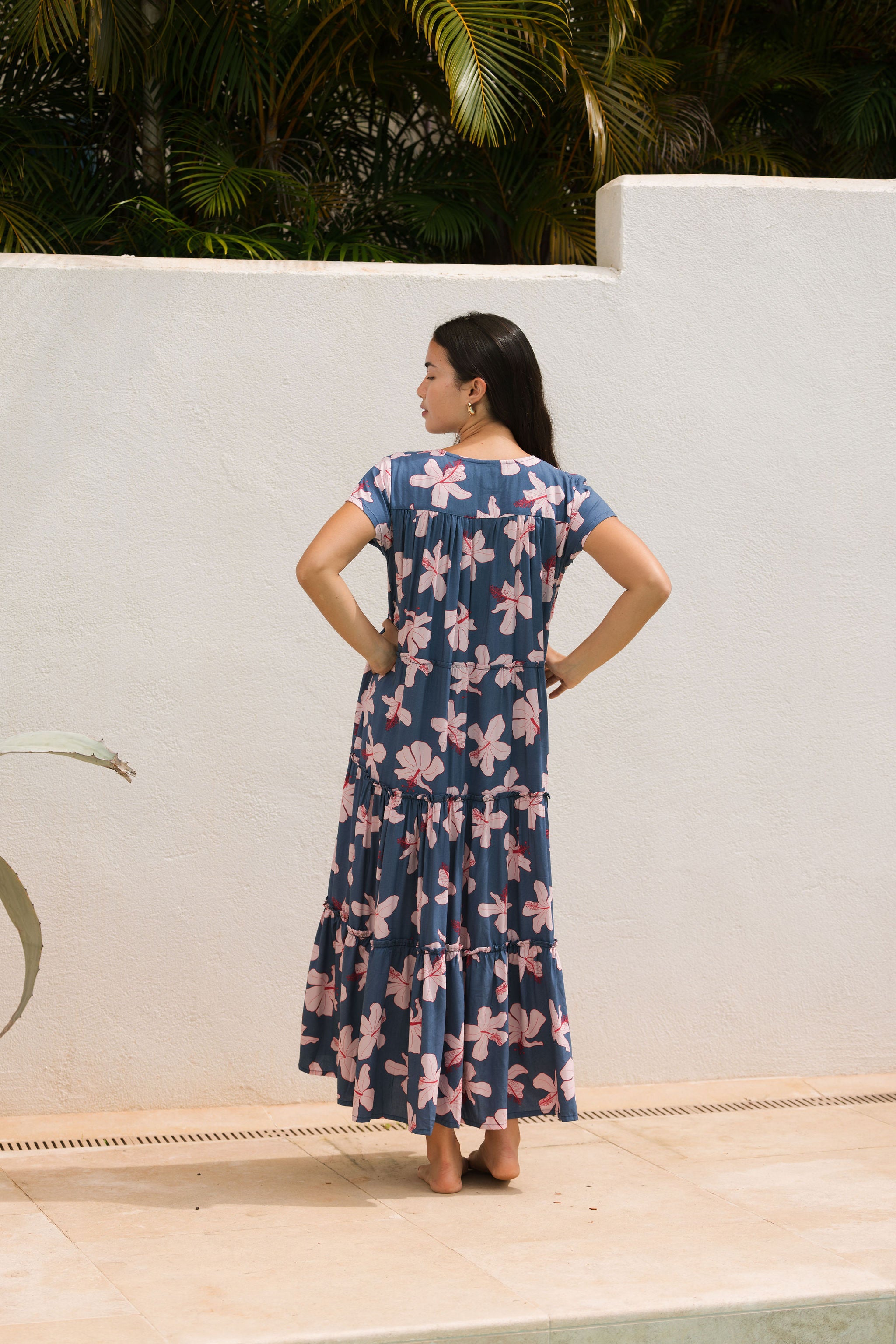 woman with black hair wearing a tiered maxi dress with v neckline in a navy and pink hibiscus print. 