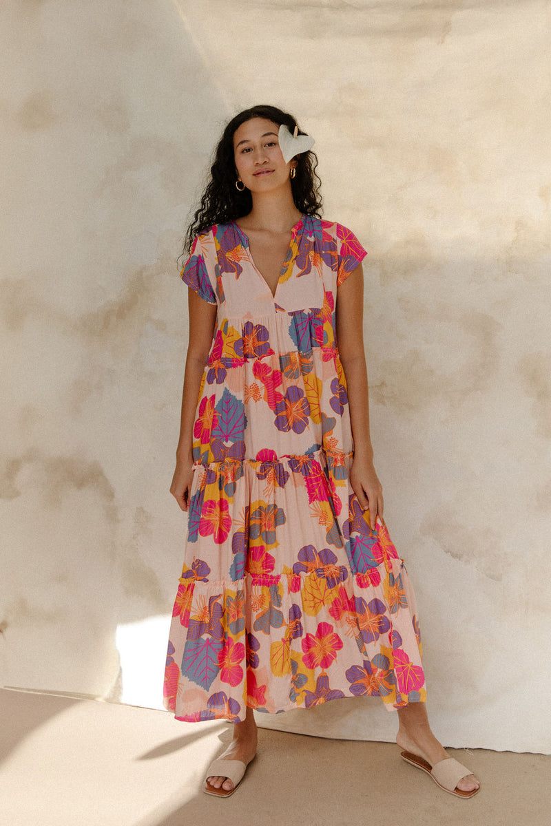 Woman in long flowy tiered dress with short sleeves and multi colored floral print with light pink ground.