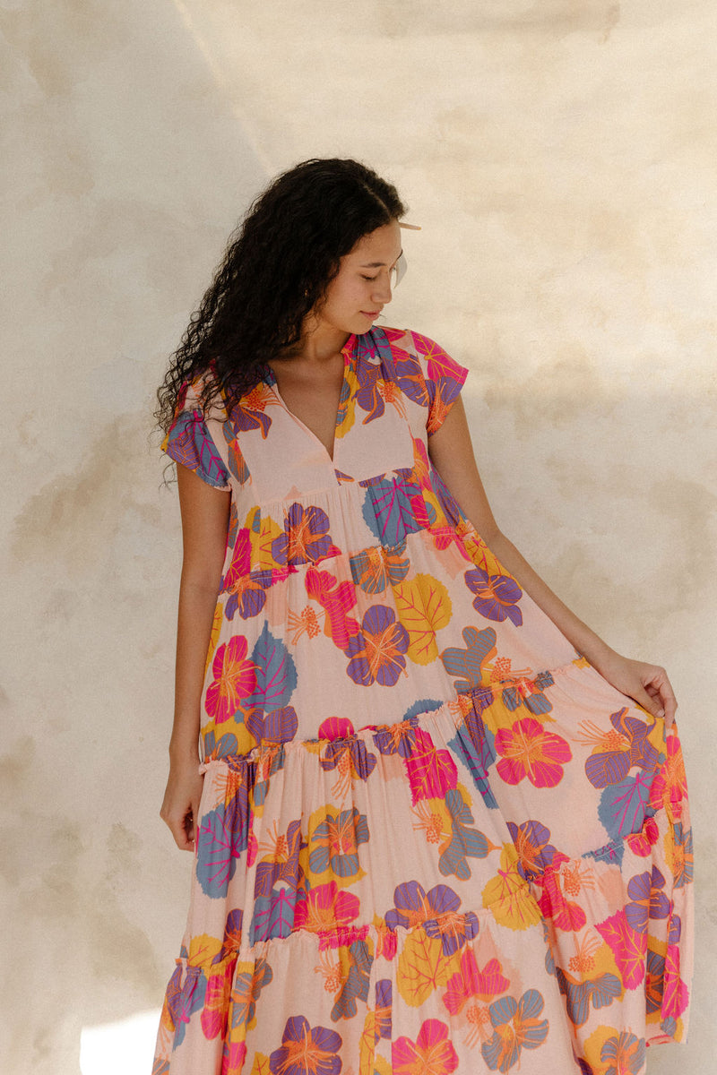 Woman in long flowy tiered dress with short sleeves and multi colored floral print with light pink ground.