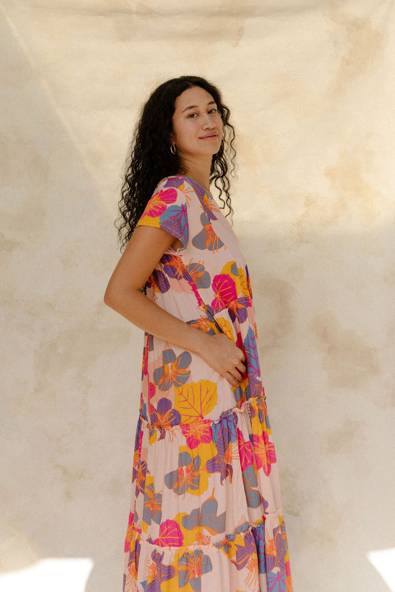 Woman in long flowy tiered dress with short sleeves and multi colored floral print with light pink ground.