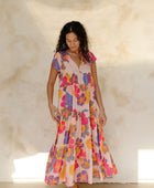 Woman in long flowy tiered dress with short sleeves and multi colored floral print with light pink ground.