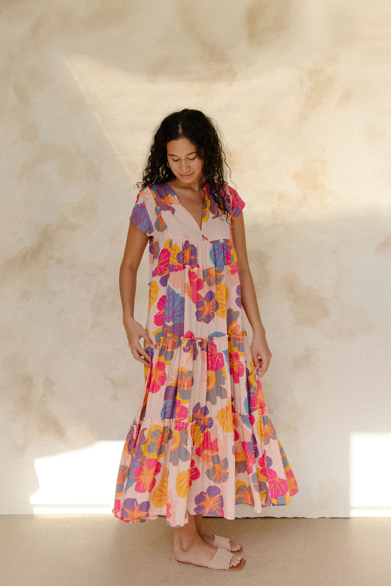 Woman in long flowy tiered dress with short sleeves and multi colored floral print with light pink ground.