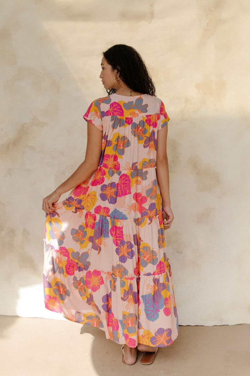 Woman in long flowy tiered dress with short sleeves and multi colored floral print with light pink ground.