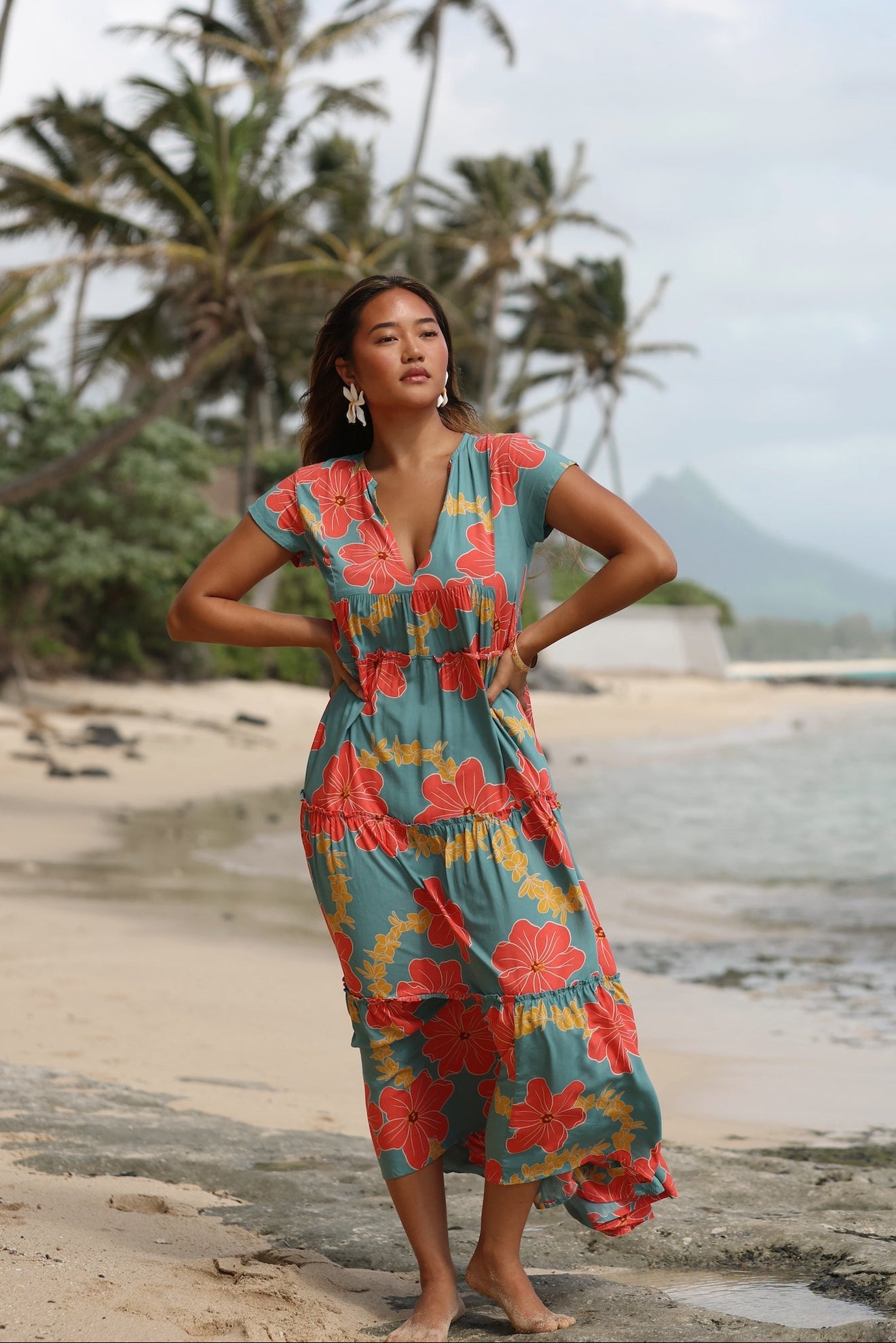 woman with brown hair wearing long flowy green maxi dress with orange and yellow lei pua print