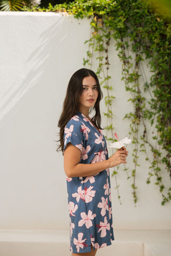 woman with black hair wearing a casual shirt dress with button up v neckline in a navy and pink hibiscus print. 