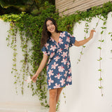woman with black hair wearing a casual shirt dress with button up v neckline in a navy and pink hibiscus print. 