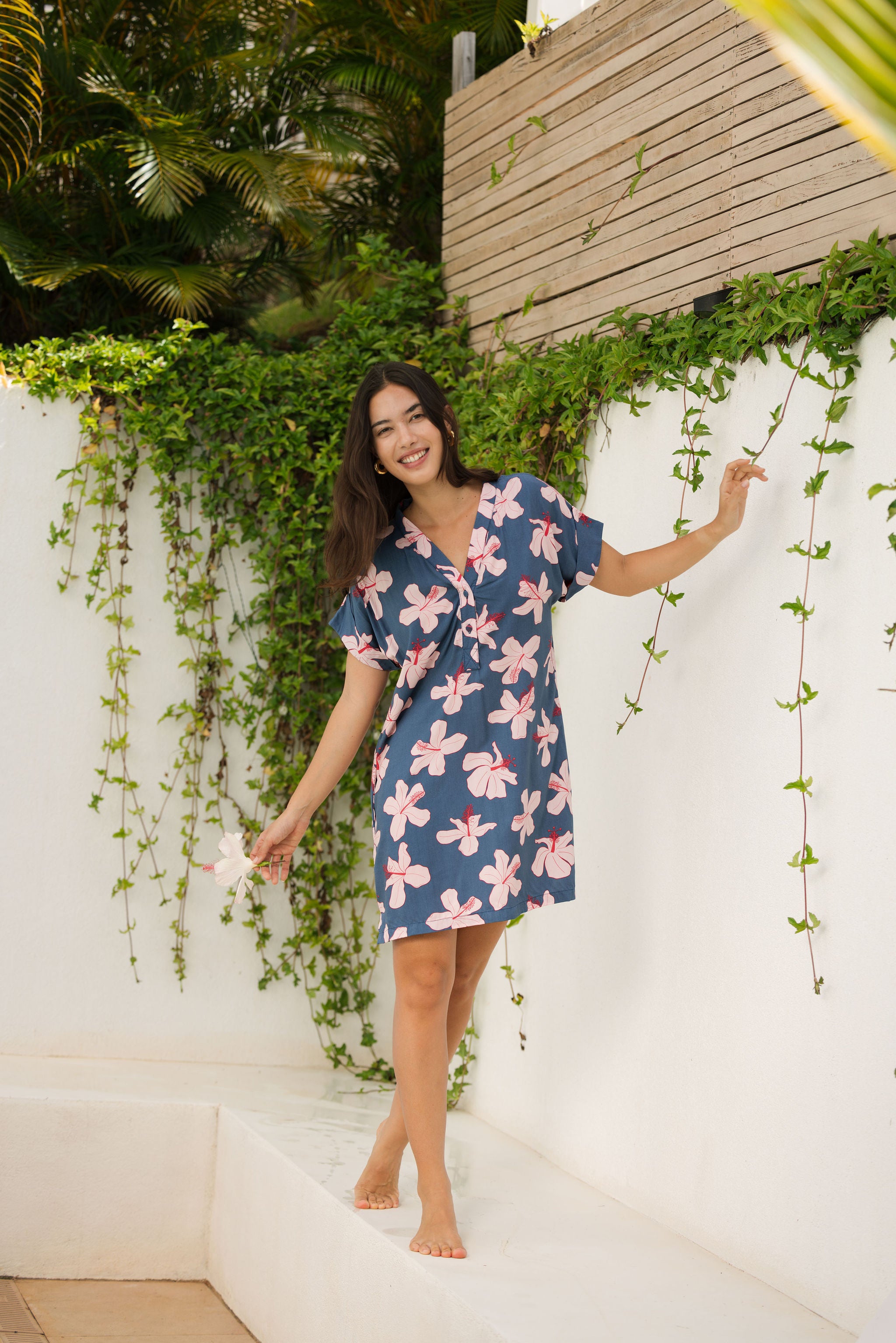 woman with black hair wearing a casual shirt dress with button up v neckline in a navy and pink hibiscus print. 