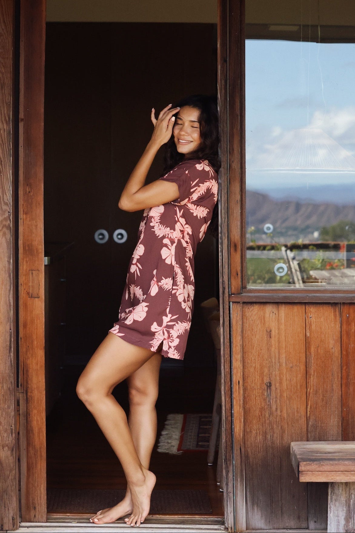 woman with brown hair wearing a brown colored button up v neckline dress with light pink lei awapuhi pattern