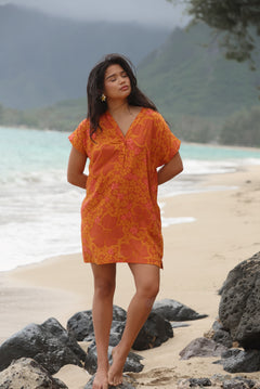woman with brown hair wearing an orange v neck short sleeve dress with puakenikeni print