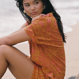 woman with brown hair wearing an orange v neck short sleeve dress with puakenikeni print