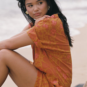 woman with brown hair wearing an orange v neck short sleeve dress with puakenikeni print