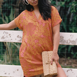 woman with brown hair wearing an orange v neck short sleeve dress with puakenikeni print