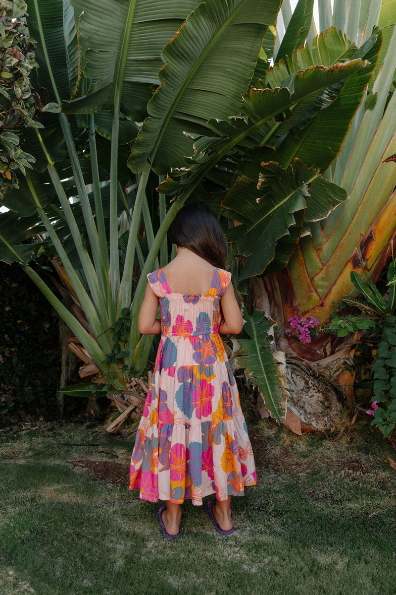 Girl wearing sleeveless long tiered dress with multi colored floral print on light pink ground.