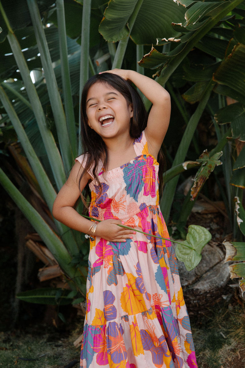 Girl wearing sleeveless long tiered dress with multi colored floral print on light pink ground.