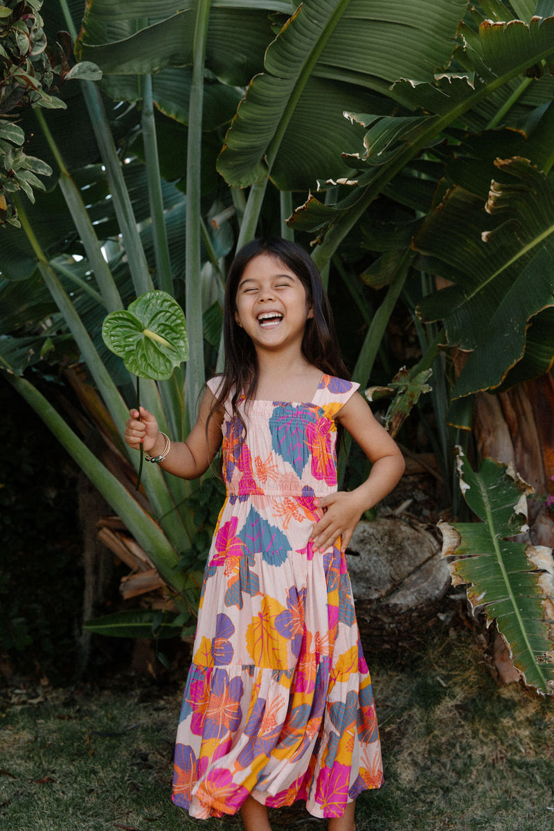 Girl wearing sleeveless long tiered dress with multi colored floral print on light pink ground.