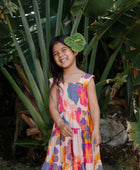 Girl wearing sleeveless long tiered dress with multi colored floral print on light pink ground.