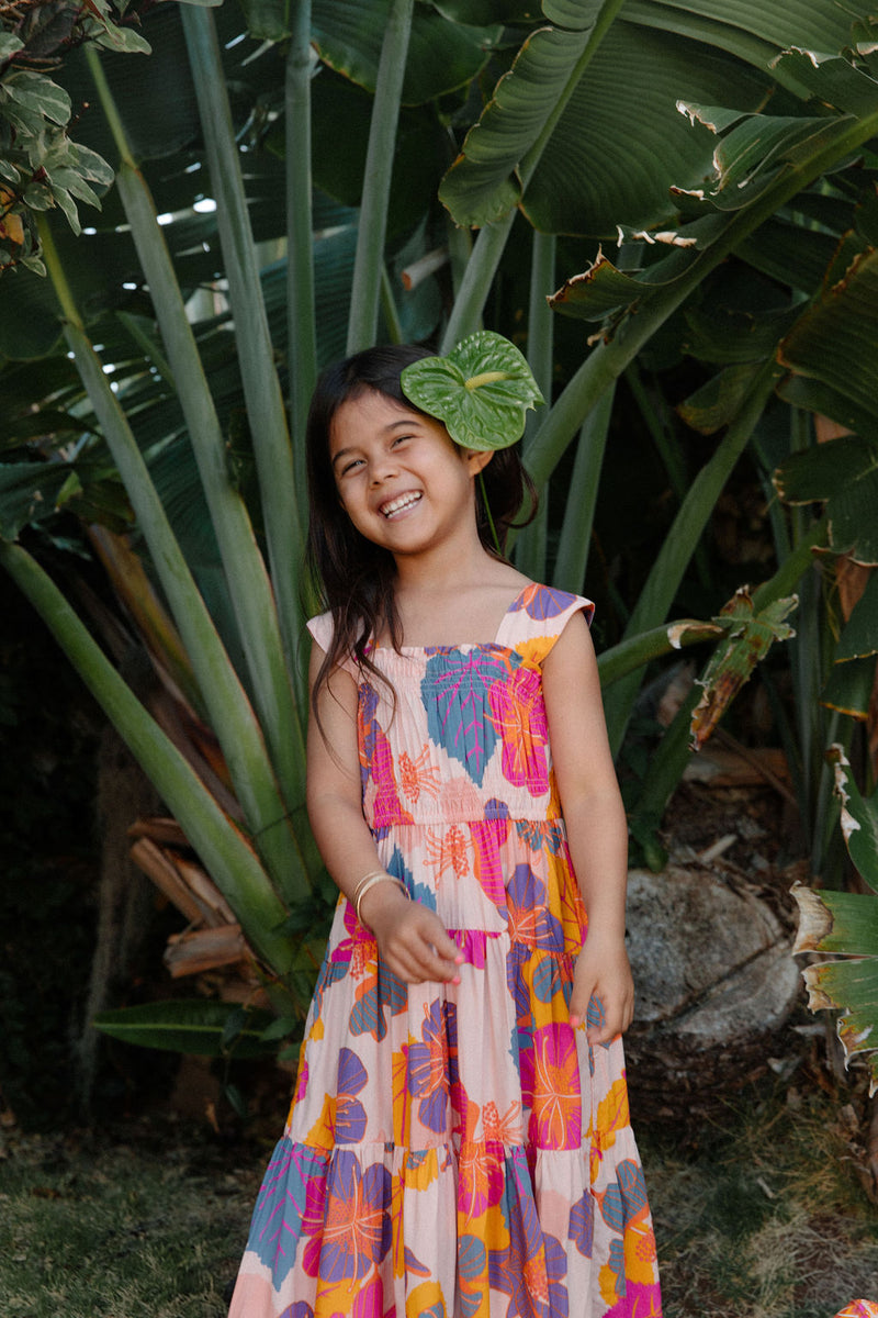 Girl wearing sleeveless long tiered dress with multi colored floral print on light pink ground.