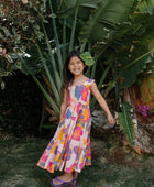 Girl wearing sleeveless long tiered dress with multi colored floral print on light pink ground.