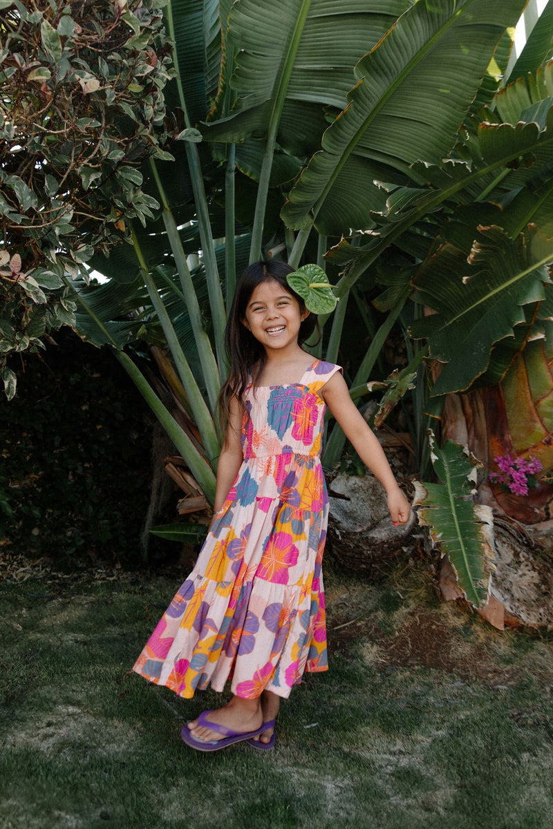 Girl wearing sleeveless long tiered dress with multi colored floral print on light pink ground.