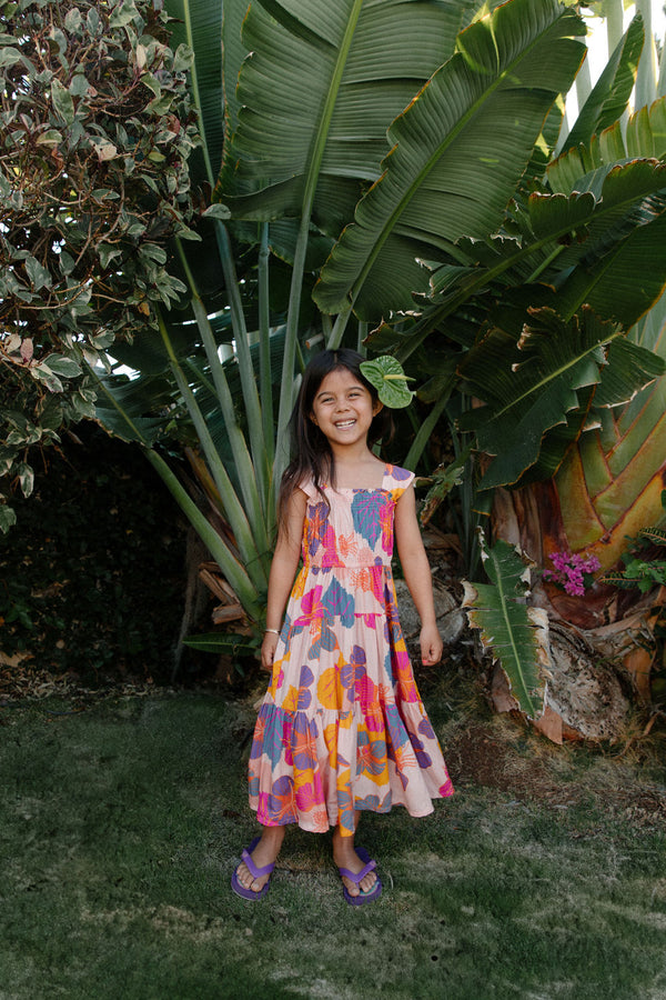 Girl wearing sleeveless long tiered dress with multi colored floral print on light pink ground.