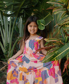 Girl wearing sleeveless long tiered dress with multi colored floral print on light pink ground.