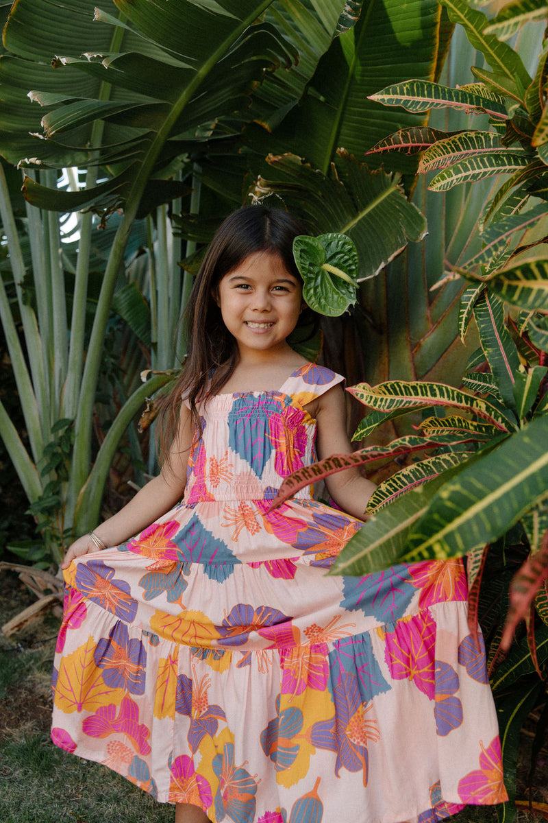 Girl wearing sleeveless long tiered dress with multi colored floral print on light pink ground.