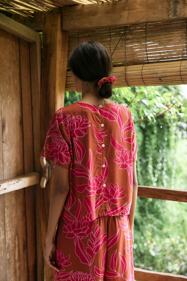 A woman with dark hair wears a YIREH t-shirt top with shell button back. It is the Noe Top in the Garden 'Awapuhi print. 