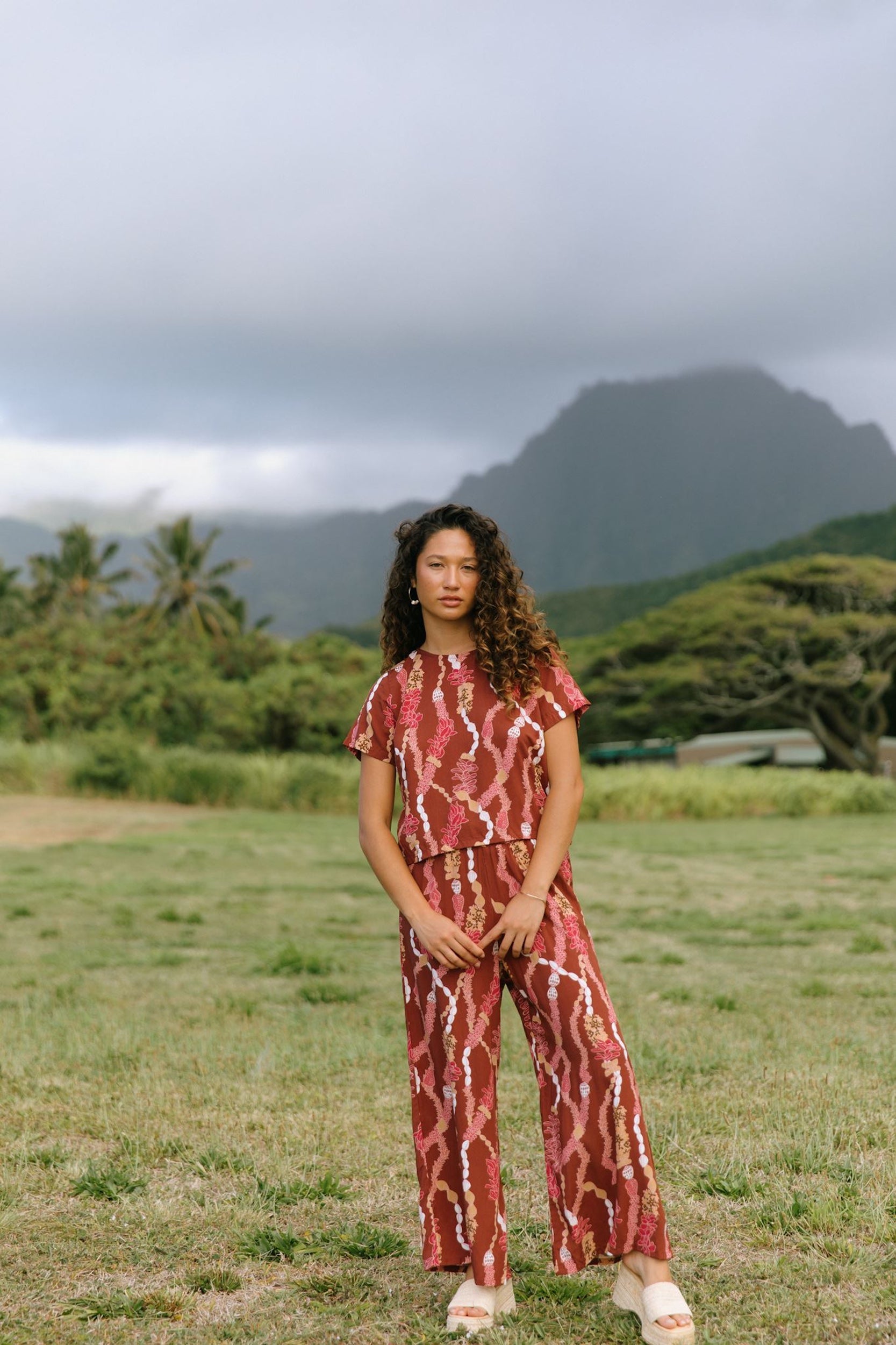 woman with brown hair wearing red-brown colored pant and top with button closure back and lei print