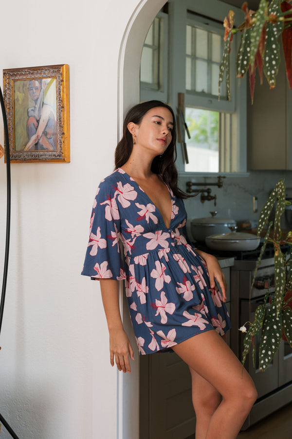 woman with black hair wearing a v neck playsuit with half sleeves in a navy and pink hibiscus print. 
